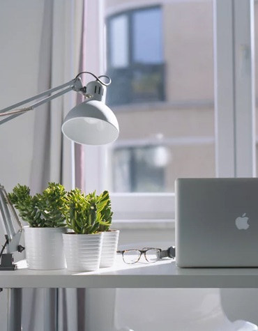 A desk by a window with reading glasses, plants and a laptop